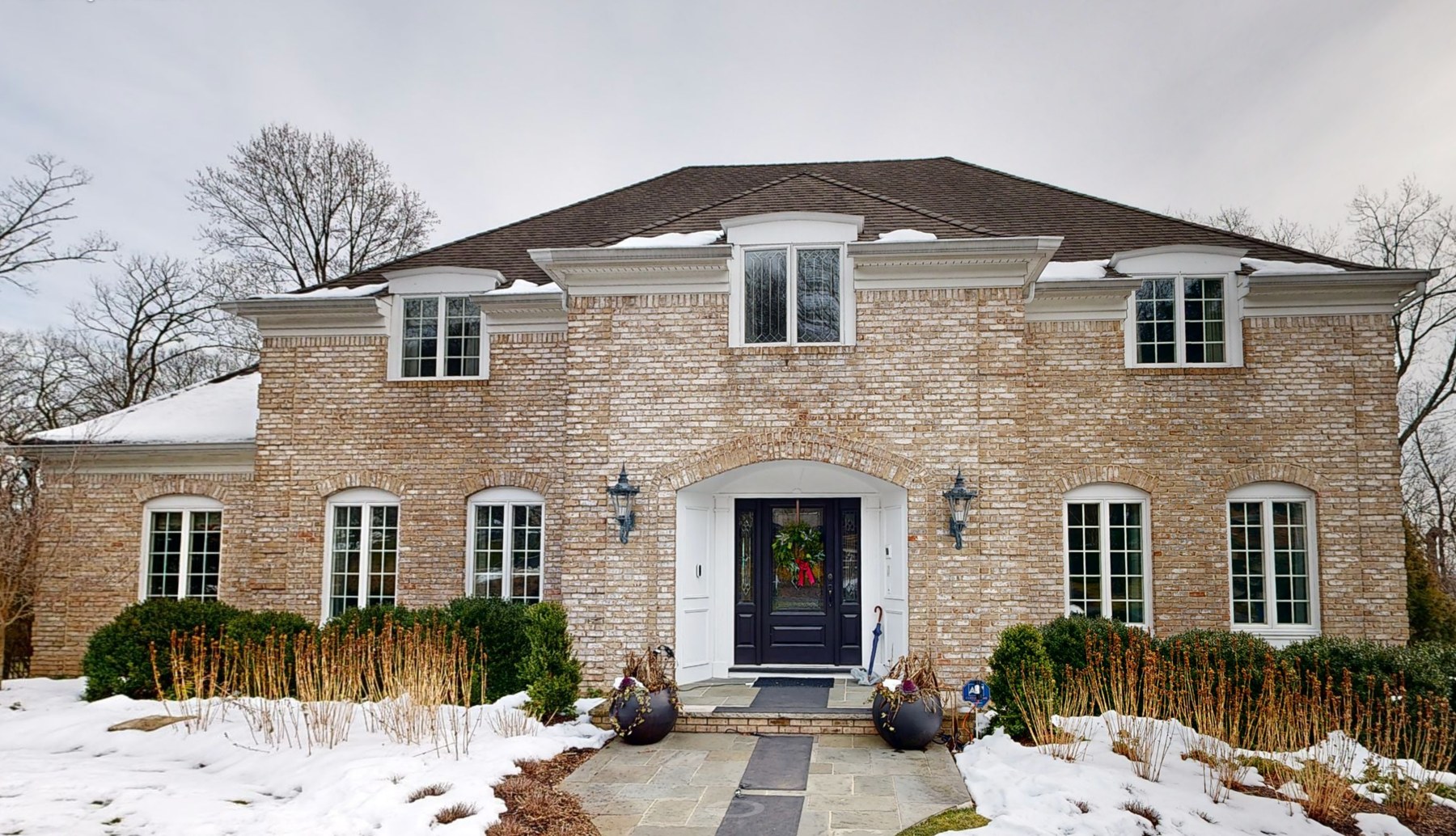 The Mountainside, New Jersey home before renovation — tan brick with an arched entry surround, a dark front door, flanking lanterns and clipped foundation planting under snow.