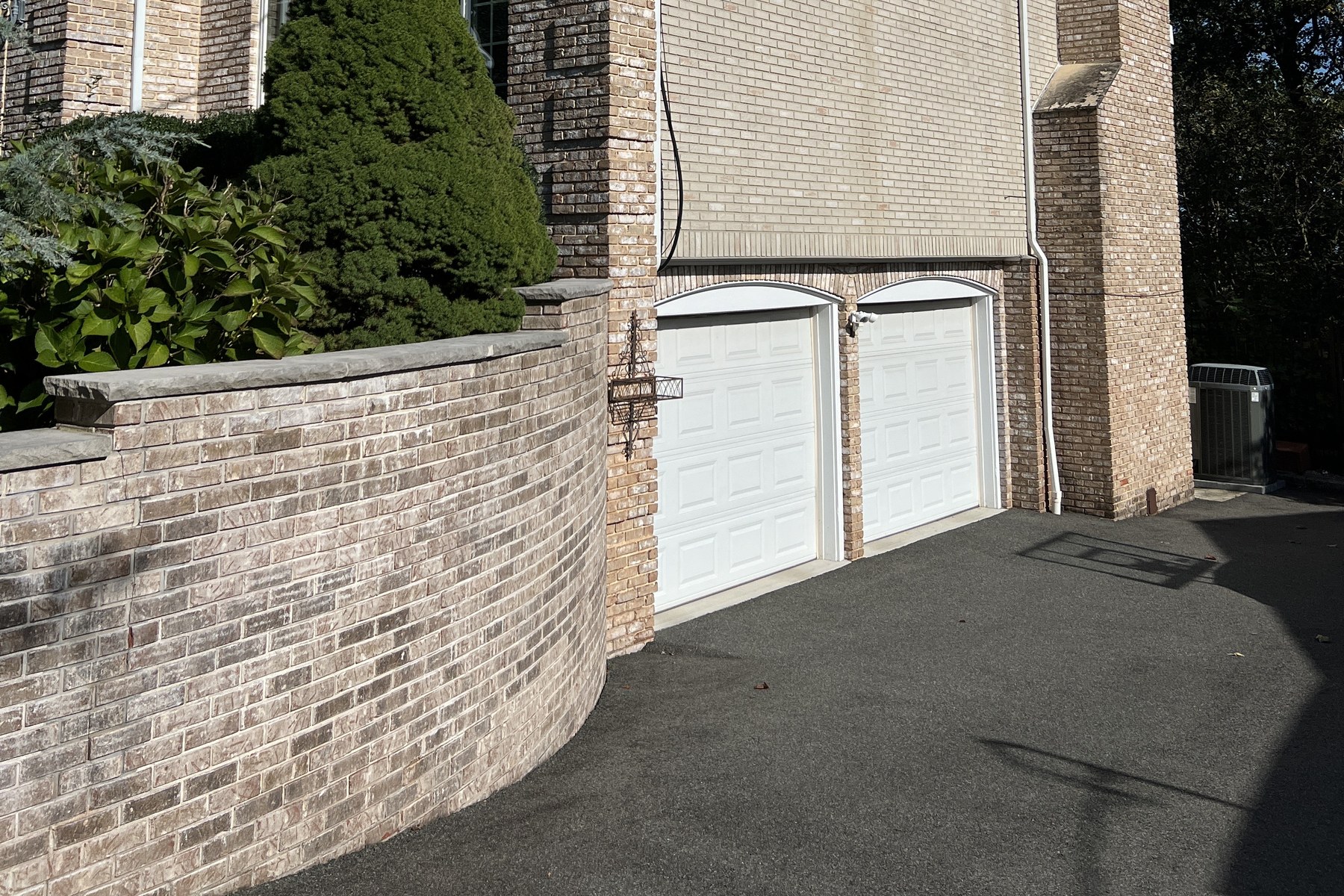 Garage and drive before renovation in Mountainside, New Jersey — tan brick with white flush-panel garage doors under a curved brick retaining wall.