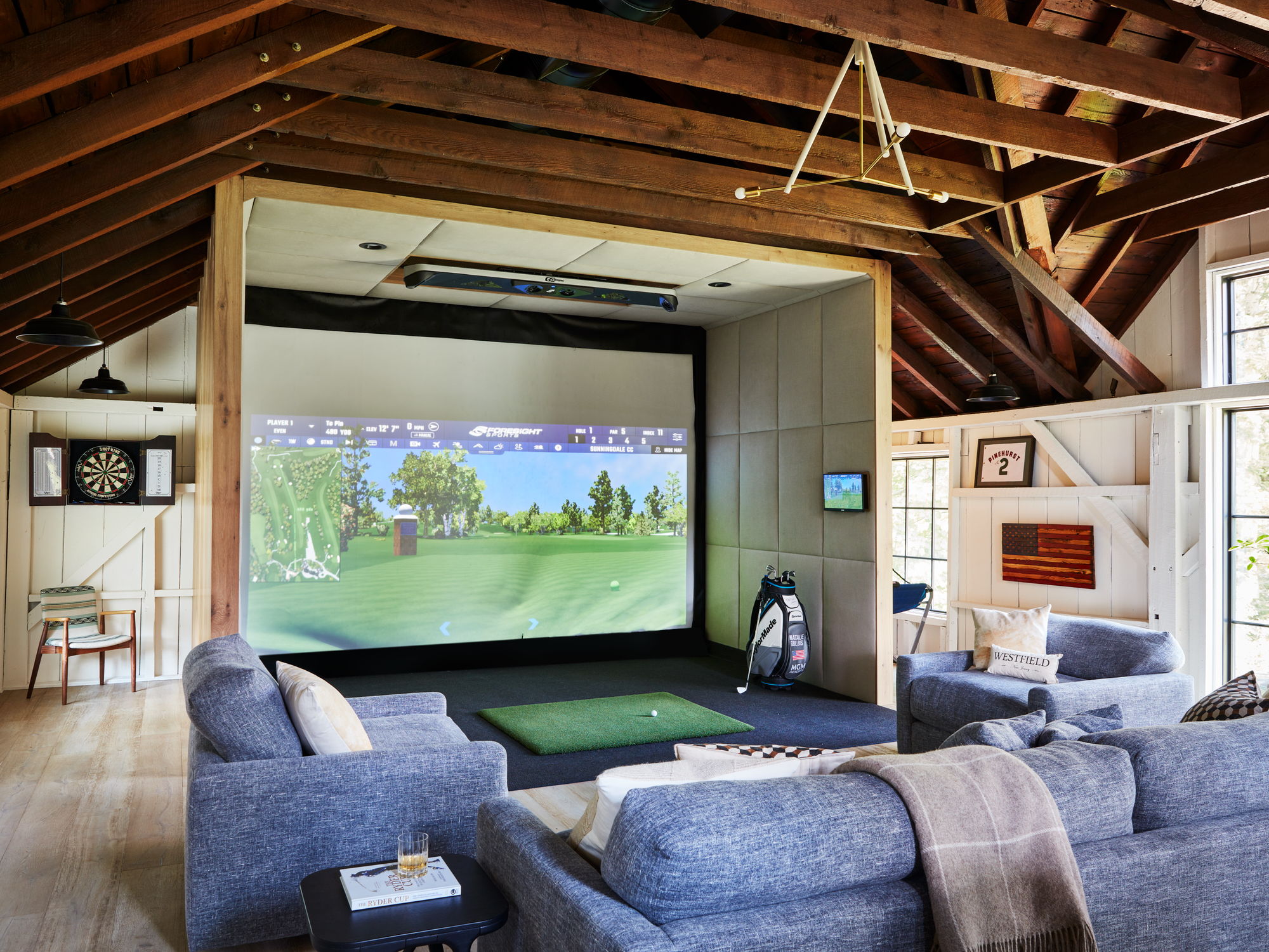 Golf simulator built into a barn with exposed rafters, a hitting mat and blue sectional seating in Westfield, New Jersey.