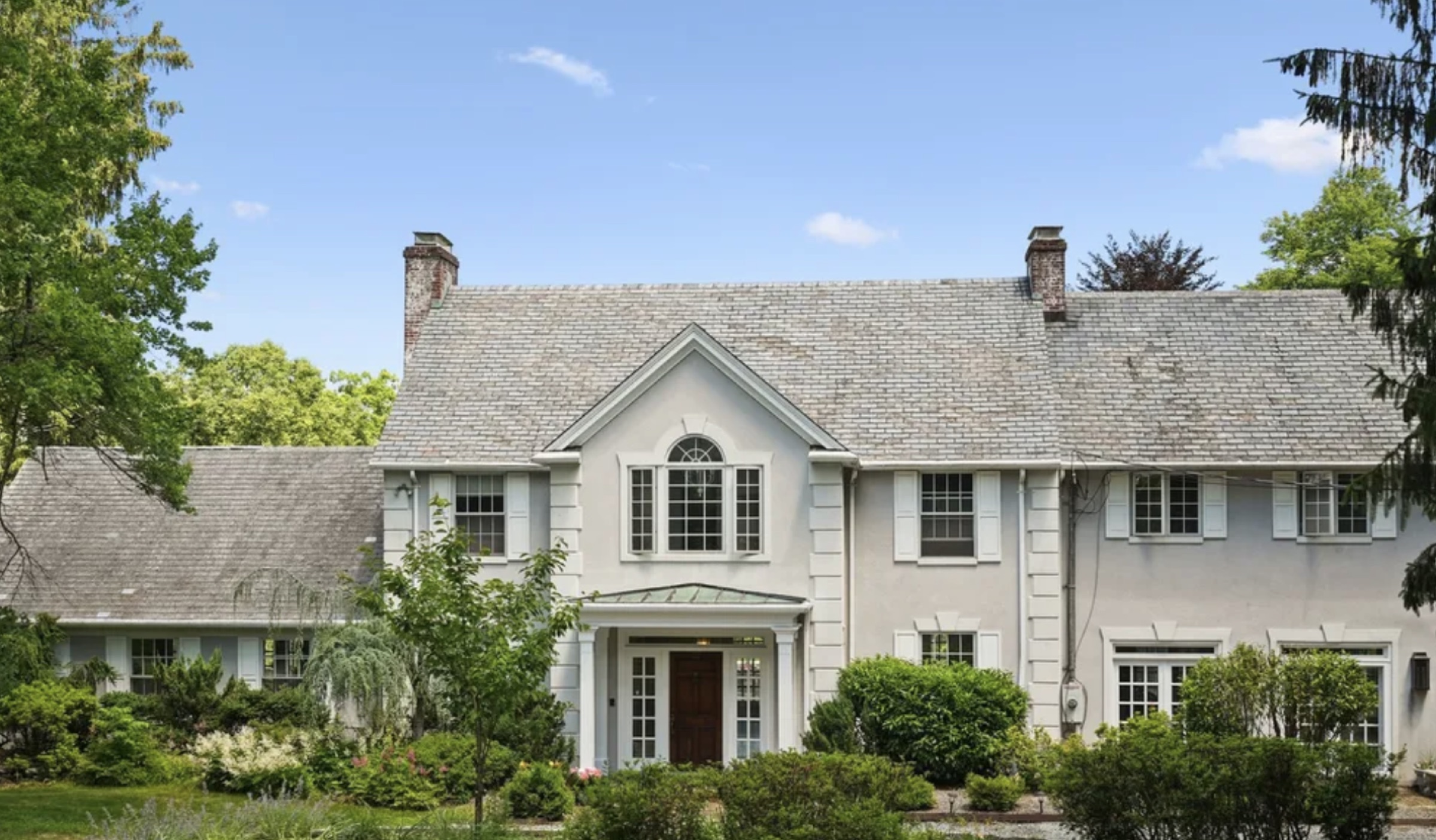The Mendham, New Jersey home before renovation — a pale stucco colonial with a weathered roof, shutters and overgrown foundation planting.