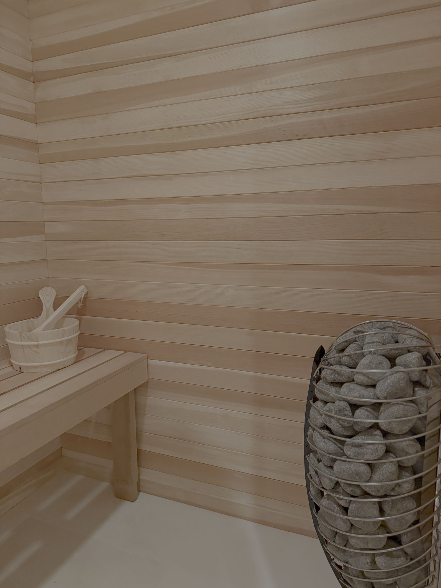 Home sauna with pale horizontal wood planking, a built-in bench and a stone-filled heater in a Short Hills, New Jersey home renovation by Ellie Mroz Design.